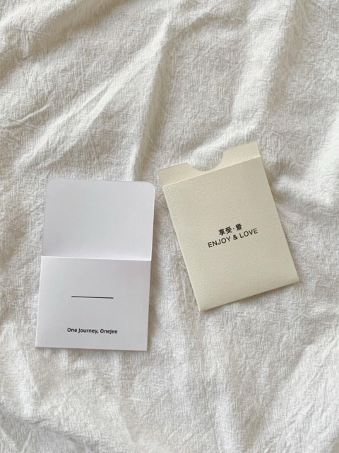 A white folded paper piece beside an ivory sleeve, arranged on softly folded white fabric.